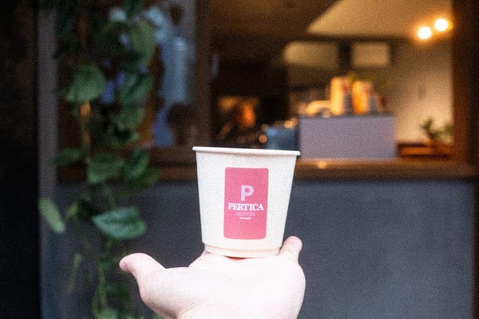 A Pertica cup held up at dusk in front of the café's service window, warm lights inside