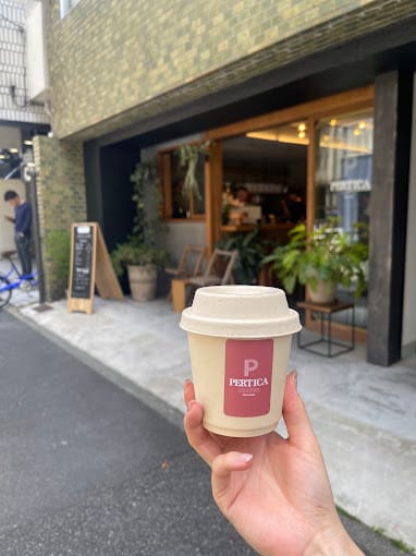 A Pertica takeaway cup with a lid, held up in front of the café's storefront on the Kodenma-chō street